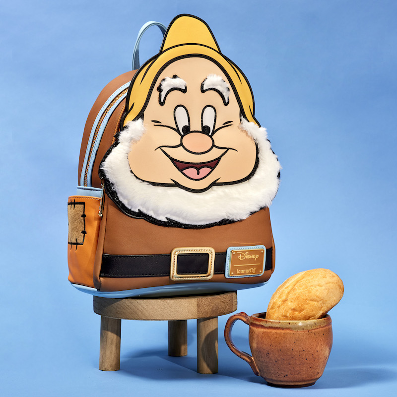 Image of the exclusive Loungefly Disney Snow White and the Seven Dwarfs Happy Mini Backpack sitting on a stool next to a mug and bread roll against a blue background